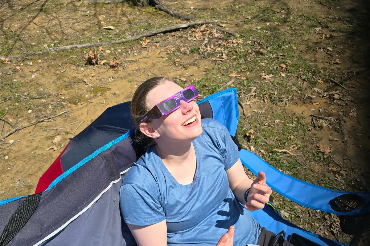 Erin reclining in a camp chair wearing purple certified eclipse glasses, looking up at the partially eclipsed sun